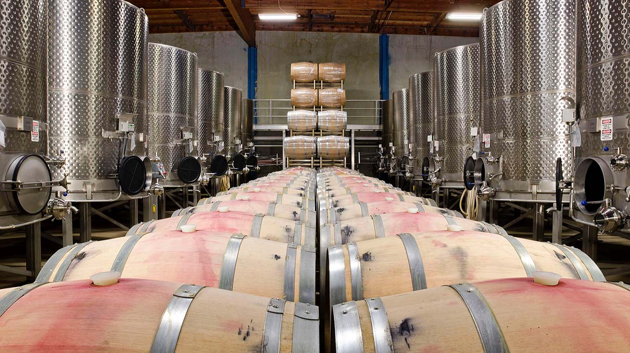 Two rows of wine stained barrels with stainless steel fermentation tanks on either side in Rodney Strong Vineyard's barrel room