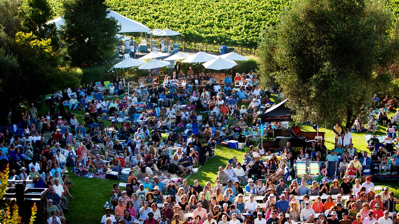 Large group of people sitting on a green lawn for a concert series with lush bright green vineyards in the background