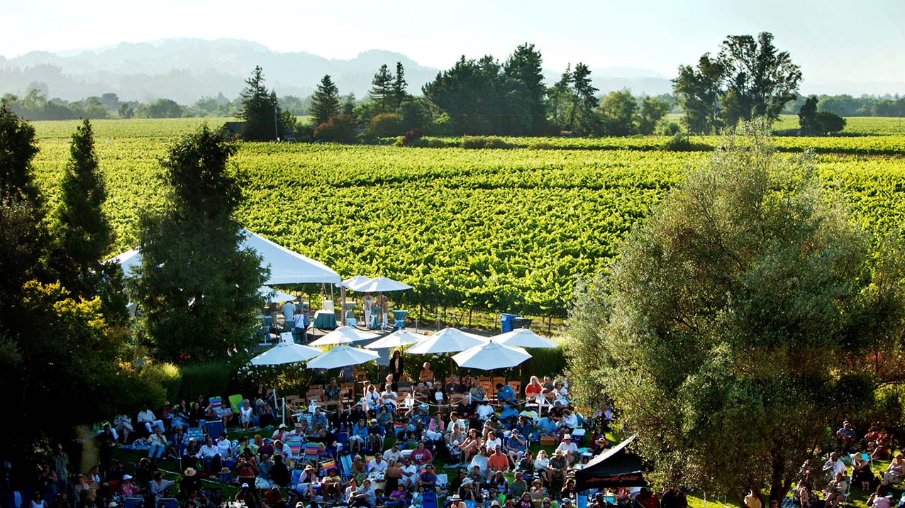 Large group of people sitting on a green lawn for a concert series with lush bright green vineyards, trees and hills in the background