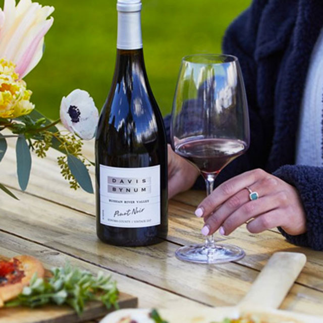 A bottle of Davis Bynum Pinot and woman's hand holding a glass of red wine while sitting at a picnic table