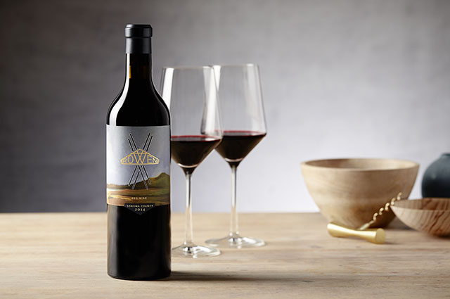 A bottle of ROWEN Red Blend, two glasses of red wine and wood bowls on a wooden table in front of a grey wall