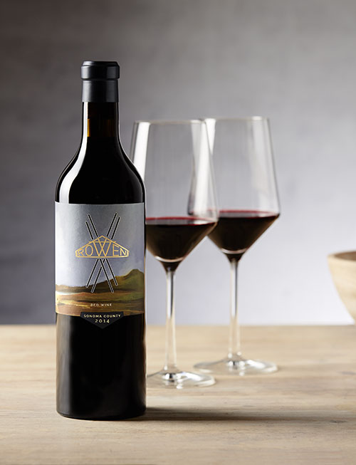 A bottle of ROWEN Red Blend, two glasses of red wine and wood bowls on a wooden table in front of a grey wall