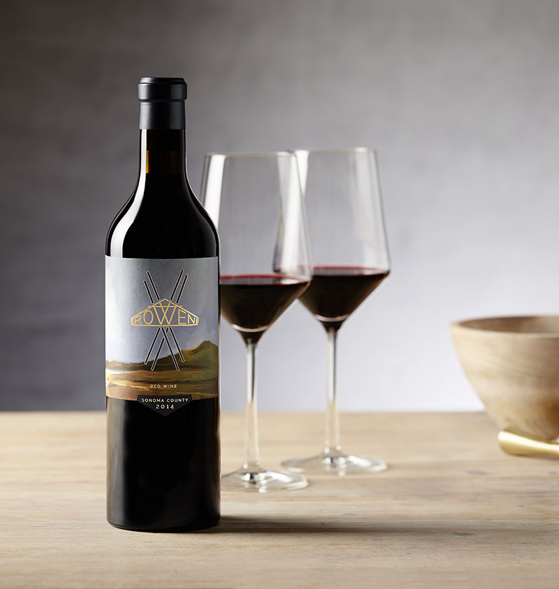 A bottle of ROWEN Red Blend, two glasses of red wine and wood bowls on a wooden table in front of a grey wall