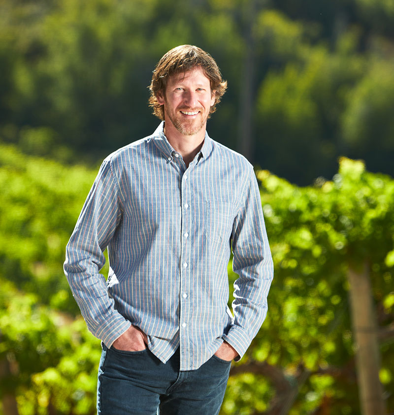Winemaker, Greg Morthole, smiles while standing in a lush green vineyard in the middle of a summer day