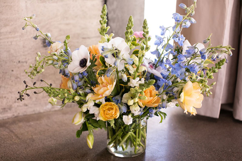 A full bouquet of spring flowers in white, yellow, pink and light blue in front of a cement wall