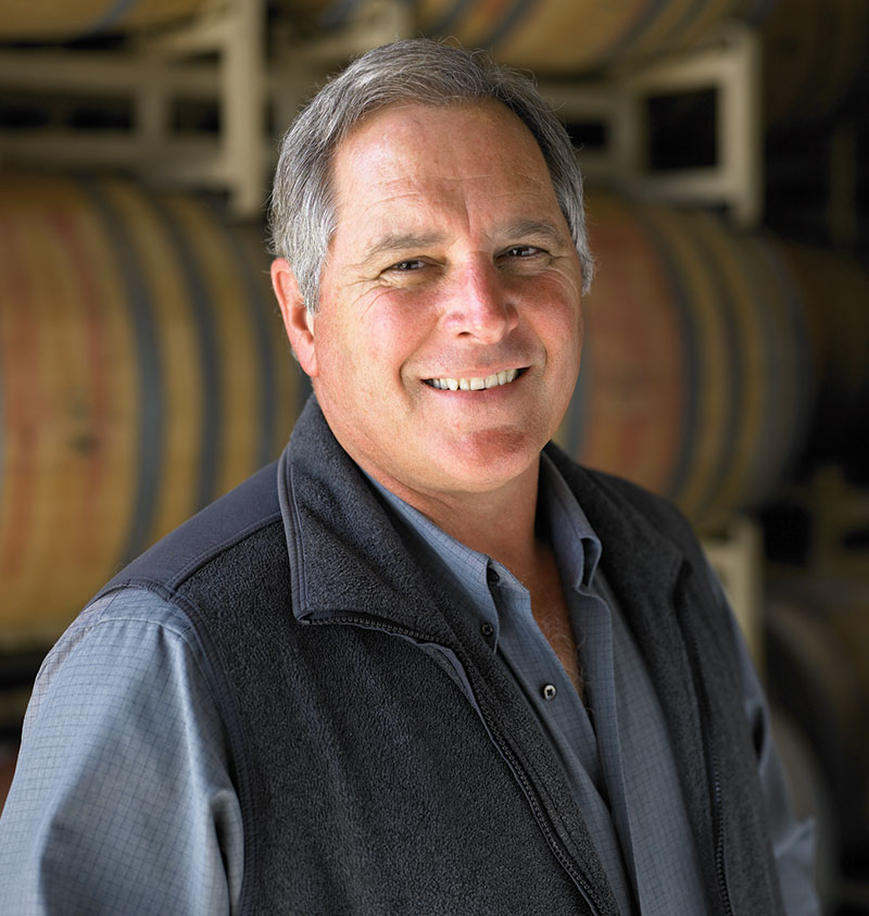 Winemaking Emeritus, Rick Sayre, smiles at the camera in front of a tall stack of wine barrels