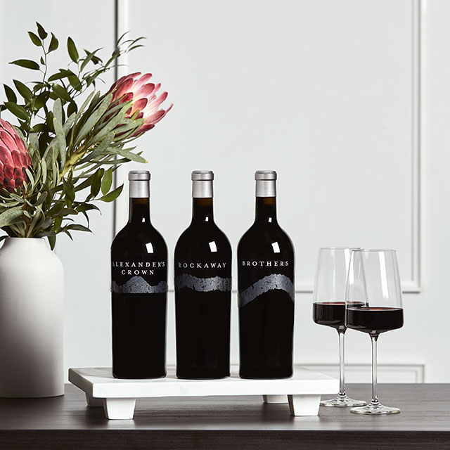 Three bottles of Rodney Strong single vineyard Cabernets on a wood table with a vase of bright pink flowers and two glasses of red wine in front of a white background