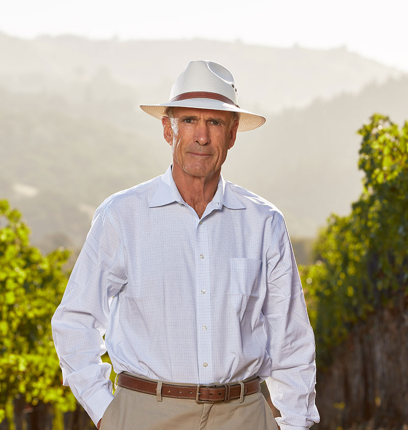 Proprietor Tom Klein stands in between two rows of a lush vineyard in the afternoon with sun hitting the hills and mountains in the background