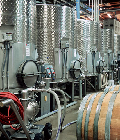Rows of tall stainless steel fermentation tanks amidst various winemaking equipment and wine barrels