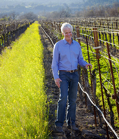 Winegrower Doug McIlroy smiles while standing in a vineyard in spring with bare vines and thick grass and mustard growing between the rows