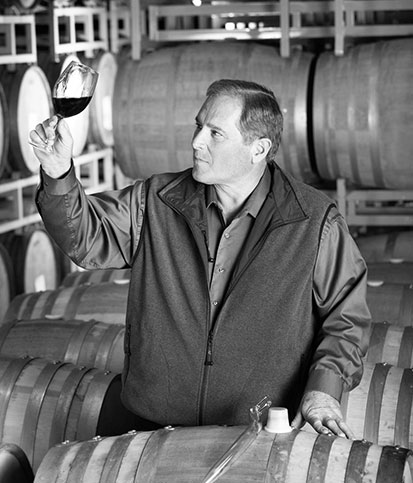 Winemaking Emeritus Rick Sayre holds up a glass of wine to evaluate while standing amidst wine barrels