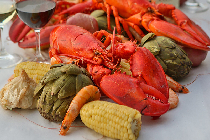 Cooked lobster, artichokes and corn on the cob spread on a white tablecloth with two glasses of wine