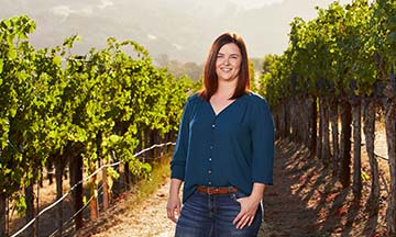 Winemaker, Olivia Wright, smiling between two rows in a green vineyard in the summer