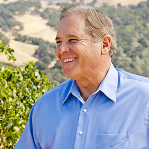 Winemaking Emeritus, Rick Sayre, smiles while looking to the distance with green vineyards and brown hills in the background