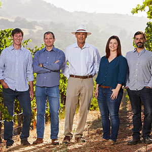 Rodney Strong Winemaking & Winegrowing team smile with Proprietor, Tom Klein, while standing in a lush green vineyard with hills and mountains in the shadows in the background