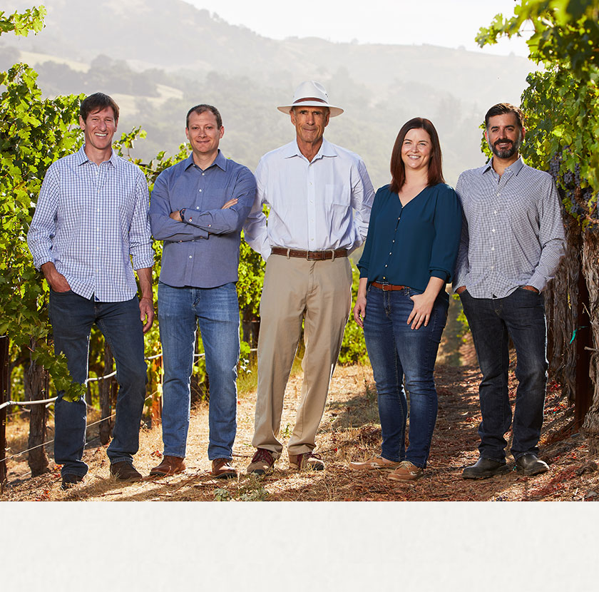 Rodney Strong Winemaking & Winegrowing team smile with Proprietor, Tom Klein, while standing in a lush green vineyard with hills and mountains in the shadows in the background