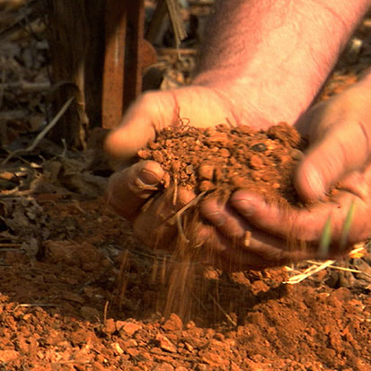 A man cups his hands in rocky red dirt while some spills out onto the ground