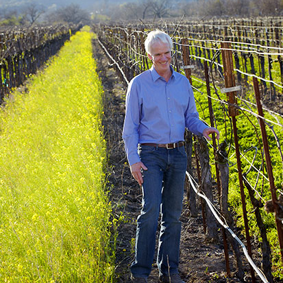 Winegrower Doug McIlroy smiles while standing in a vineyard in spring with bare vines and thick grass and mustard growing between the rows