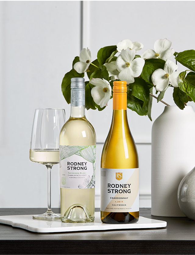 Two bottles of Rodney Strong wine sit on a table in front of a white wall.