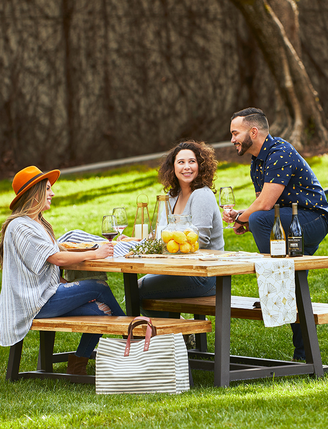 A trio of guests sit smiling at a picnic table on the Green at Rodney Strong Vineyards while they drink wine.