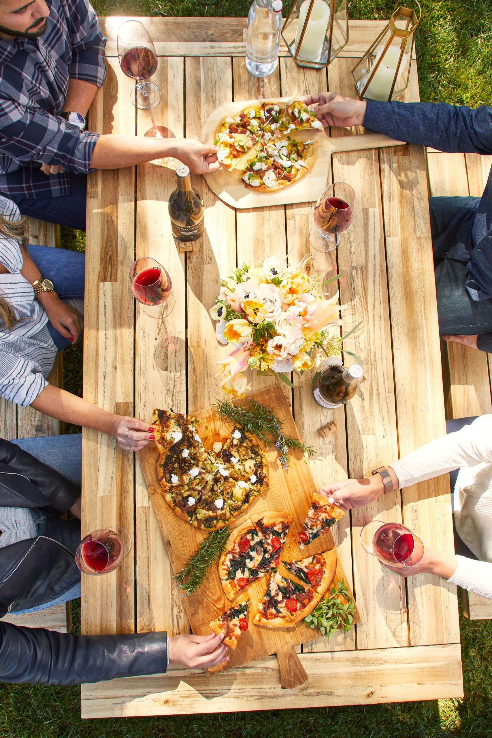 Several arms reach across a light wood picnic table laden with glasses of wine, fire roasted pizzas, lanterns and flowers in the afternoon light