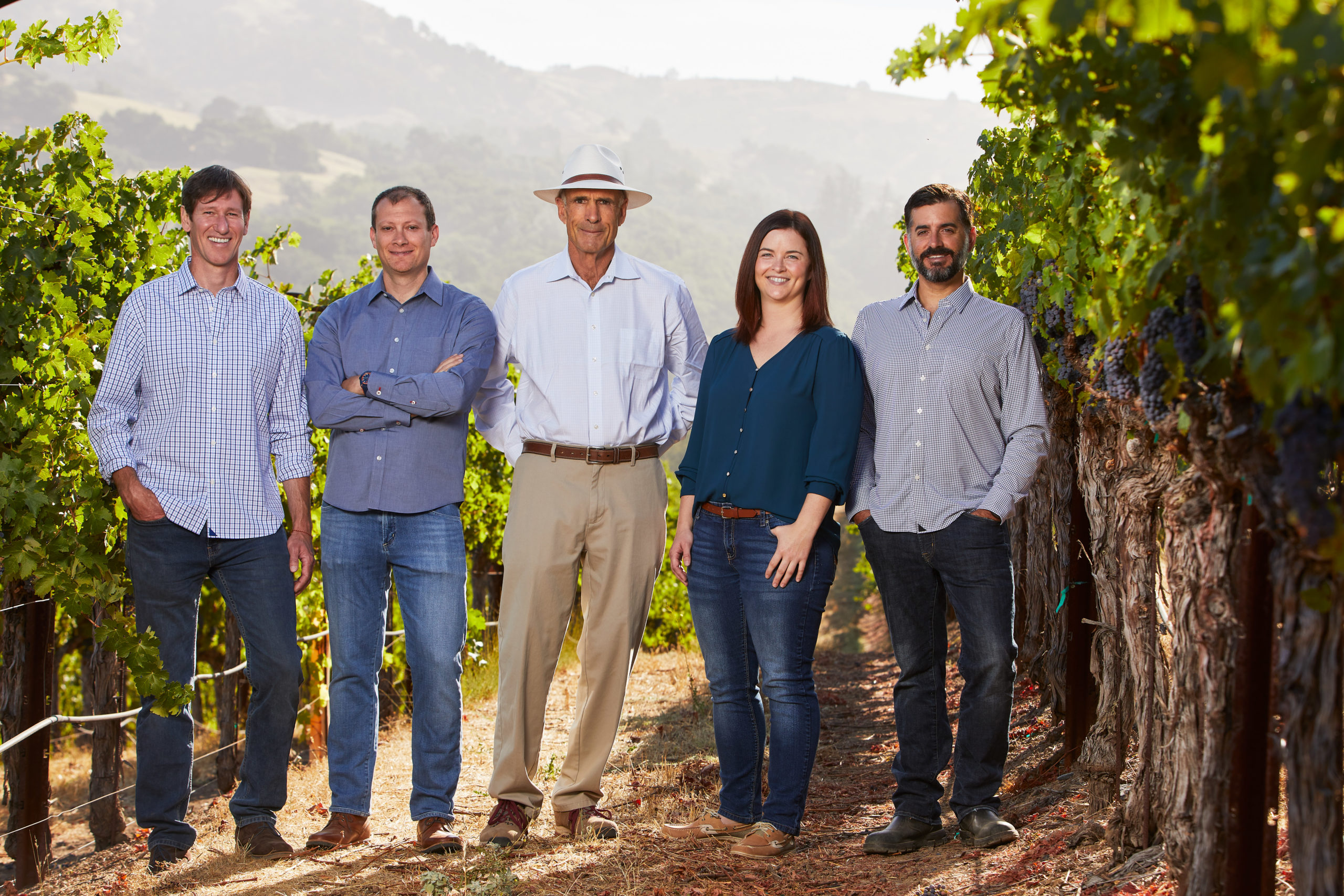 Four men and one woman stand smiling in a lush green vineyard with the setting sun behind them