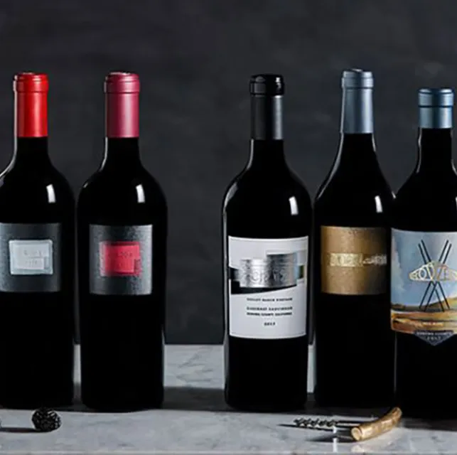 Five bottles of ROWEN Wine Company wines on a marble table with a glass of wine, vase of white flowers and assorted fruits in front of a dark grey wall