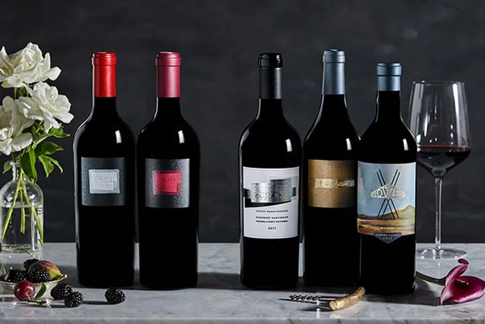 Five bottles of ROWEN Wine Company wines on a marble table with a glass of wine, vase of white flowers and assorted fruits in front of a dark grey wall