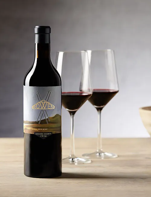 A bottle of ROWEN Red Blend, two glasses of red wine and wood bowls on a wooden table in front of a grey wall