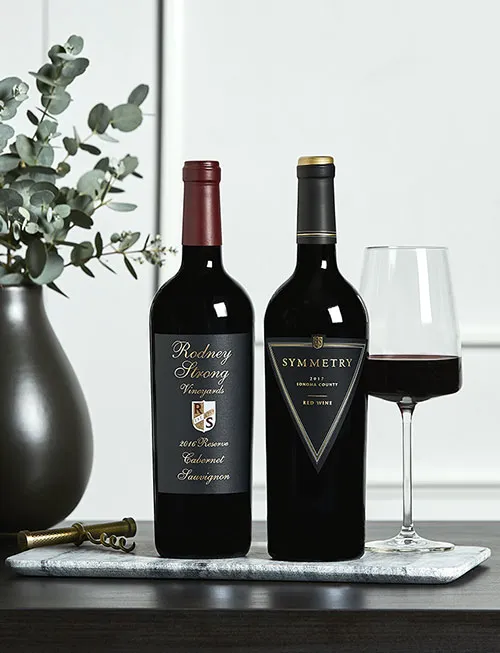 Two bottles of Rodney Strong red wine on a wood table with vases of dried flowers in front of a white background